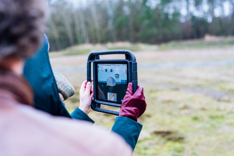 Führung dem Gelände der Gedenkstätte Bergen-Belsen, eine Person hält ein digitales Tablet mit Informationen zur Gebäudestruktur in der Hand