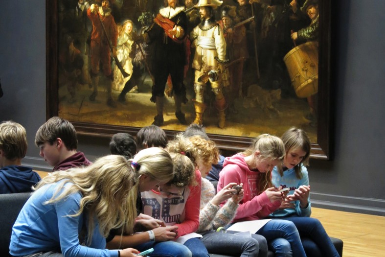 Mehrere Schüler*innen sitzen vor Rembrandts Werk "Nachtwache", schauen aber auf ihre Smartphones