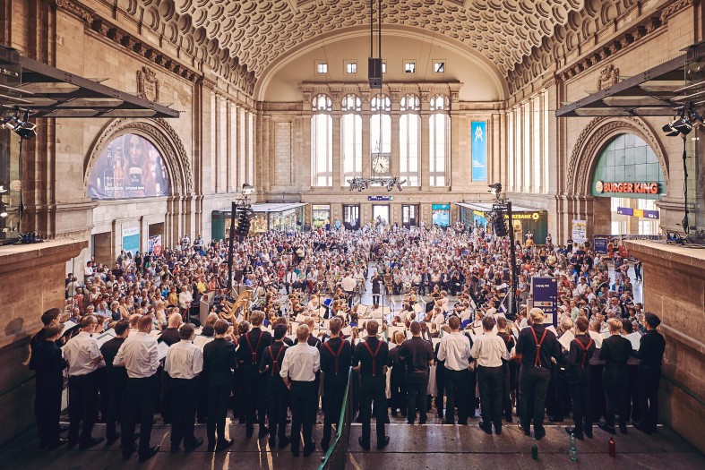 Hunderte Menschen sitzen in einem Bahnhofsgebäude, vor ihnen ein Chor samt Musiker*innen