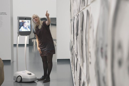 Eine Roboter auf dem ein Display installiert ist, steht neben einer Kunstvermittlerin. Das Display oder Tablett zeigt eine Ausstellungsbesucherin, die sich via Livestream und mithilfe des Roboters nun durch die Ausstellung bewegen kann.